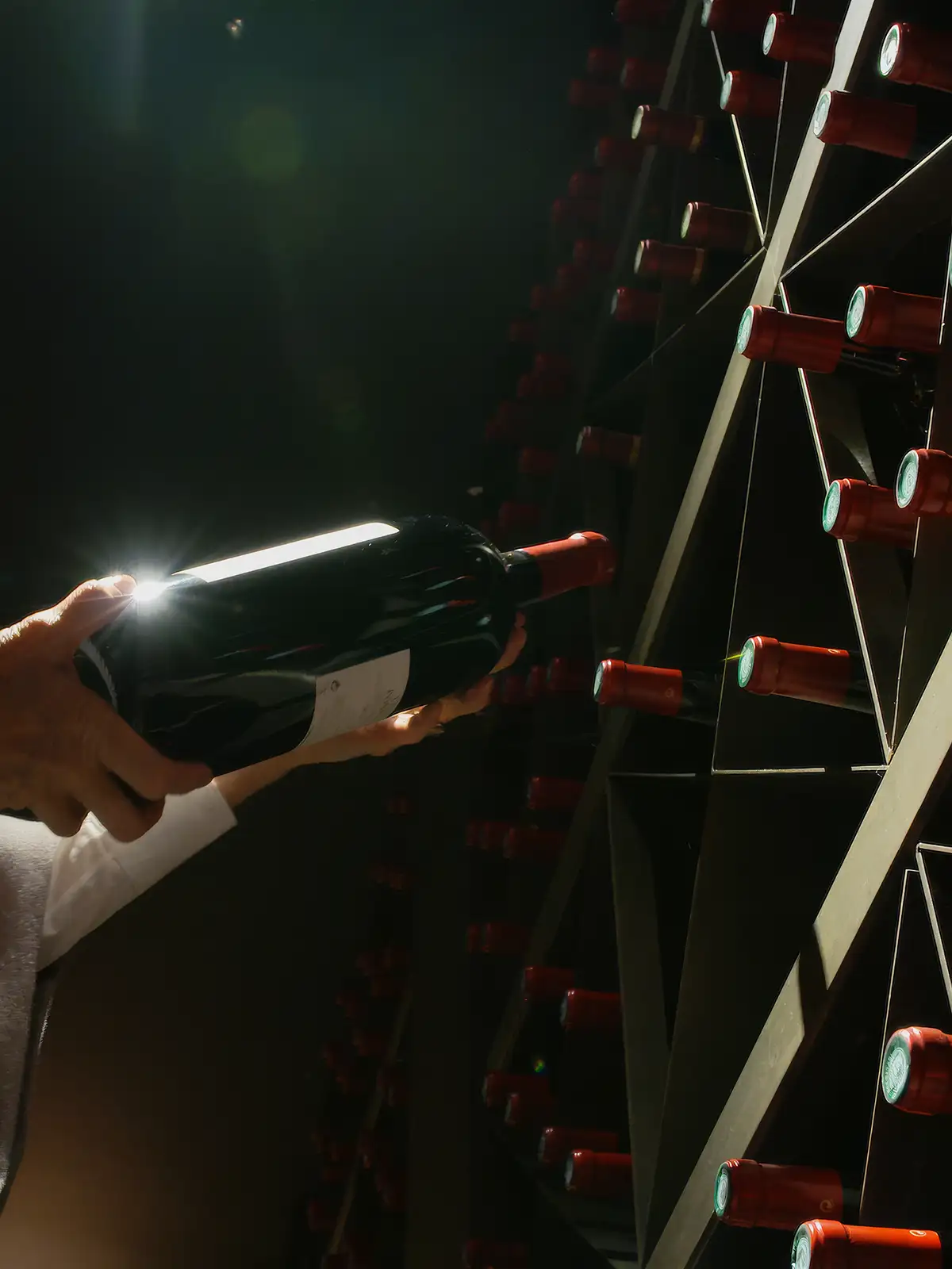 A person pulling a dark wine bottle from a triangular wooden cellar rack.