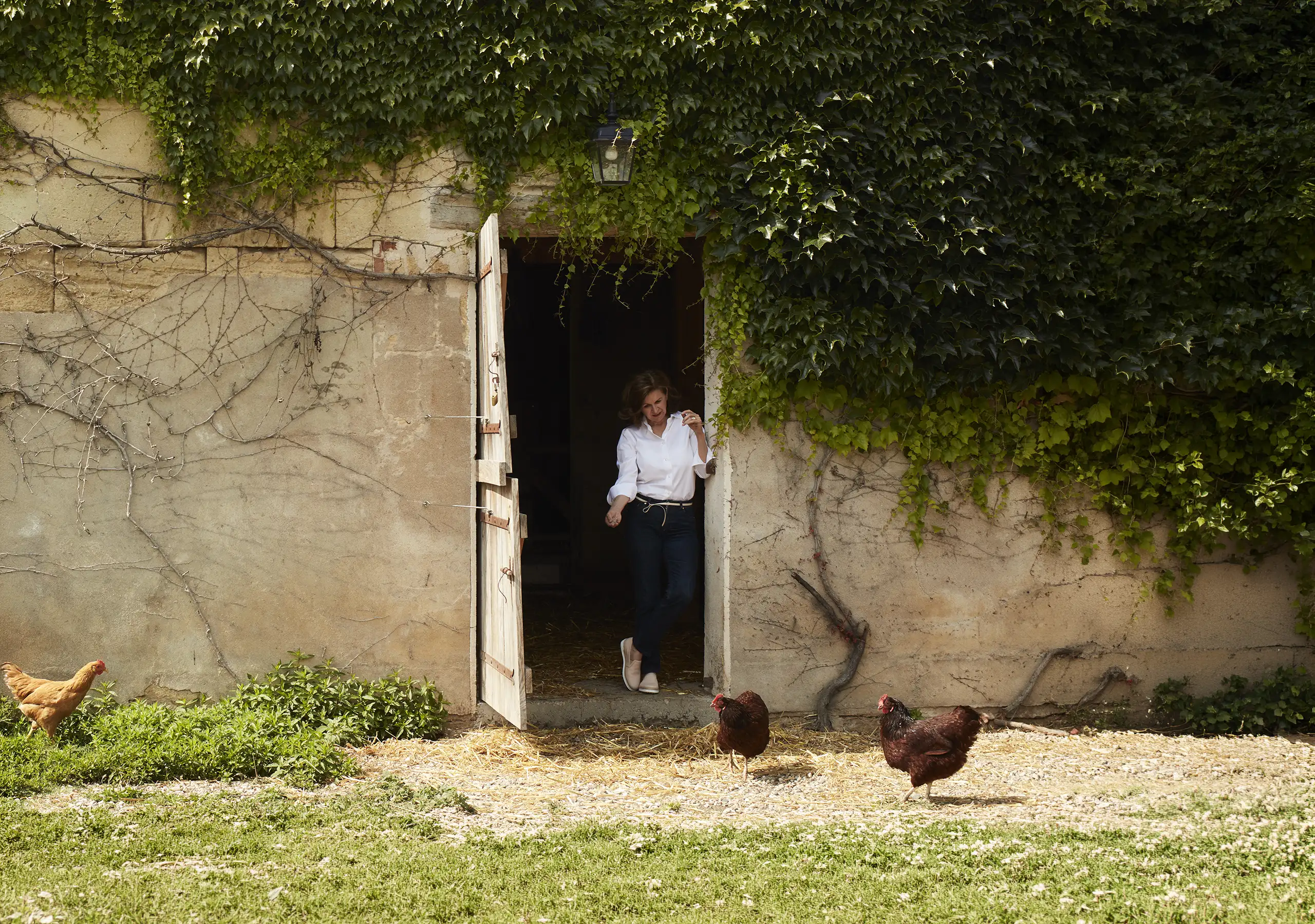 A woman stands in the doorway of an ivy-covered stone building with three chickens in the foreground.